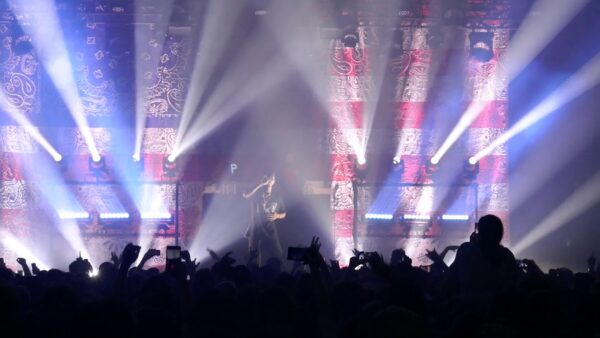Joey Bada$$ performing in front of an American flag print backdrop with white lights shining into a full audience.